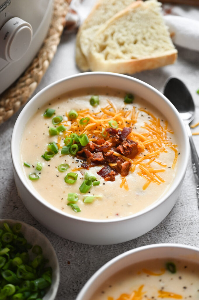 A bowl of slow cooker potato soup topped with bacon, green onions and cheddar cheese with a couple pieces of bread next to it.