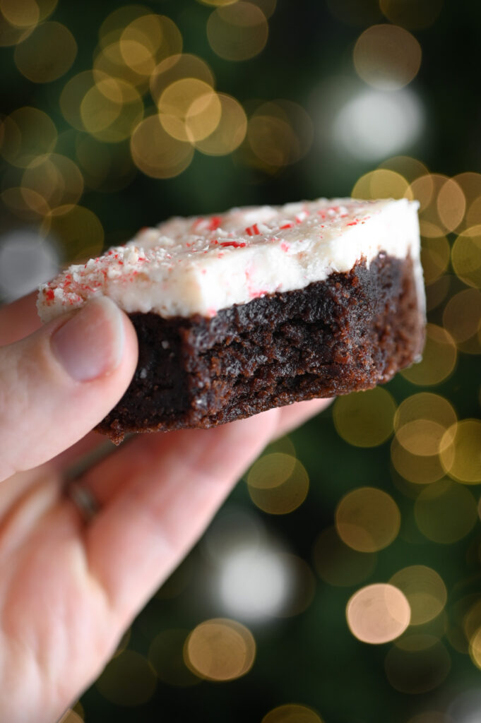 A hand holding a peppermint bark brownie with a bite out of it in front of a lit Christmas tree.
