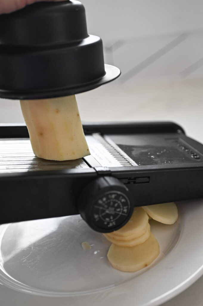 A peeled russet potato being sliced on a black mandoline.