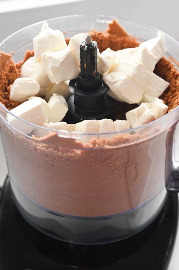 Pulverized Biscoff cookies in a food processor with cubes of cream cheese on top.