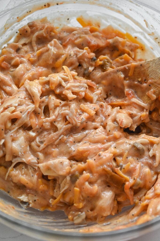 A spoon stirring a mixture of soups, chicken and cheese in a clear bowl.