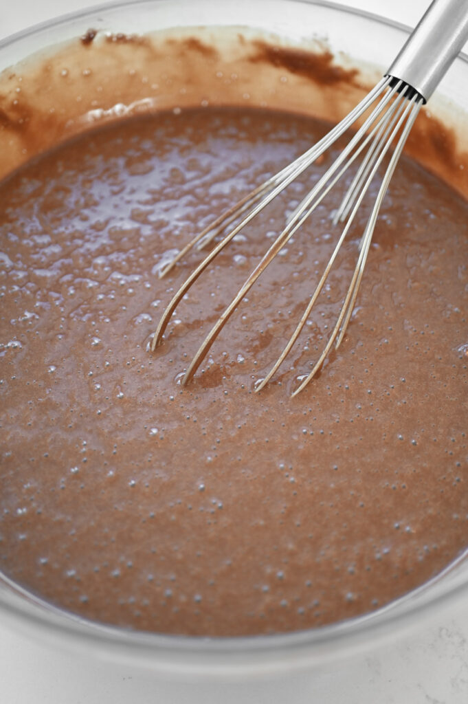 A whisk stirring chocolate cupcake batter in a large clear bowl.
