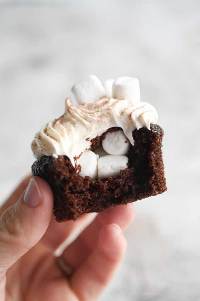 A hand holding a cupcake with a bite out of it showing the center filled with mini marshmallows.