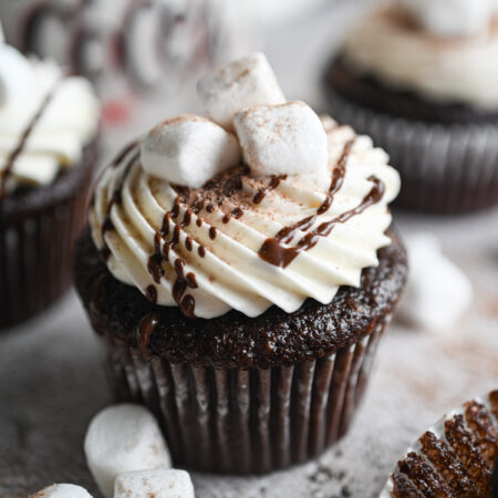 A hot chocolate cupcake with frosting, a drizzle of chocolate and a few mini marshmallows on top.
