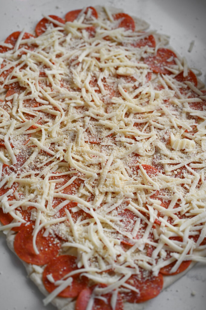 A sheet of dough topped with pepperoni, cheeses and seasonings.