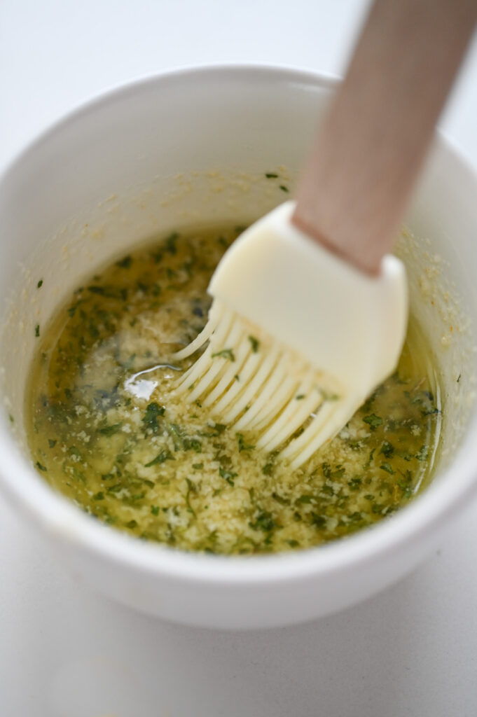 A pastry brush stirring together melted butter, fresh parsley, parmesan cheese and garlic powder.