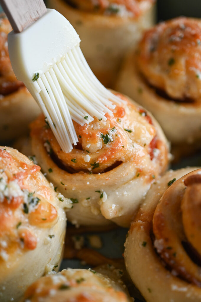 A pastry brush adding melted butter with garlic and parsley over some cheesy baked rolls.