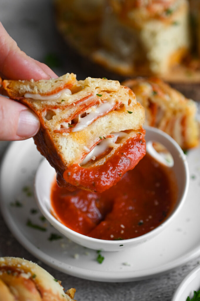 Half of a pepperoni roll being dipped into a small white bowl of pizza sauce.