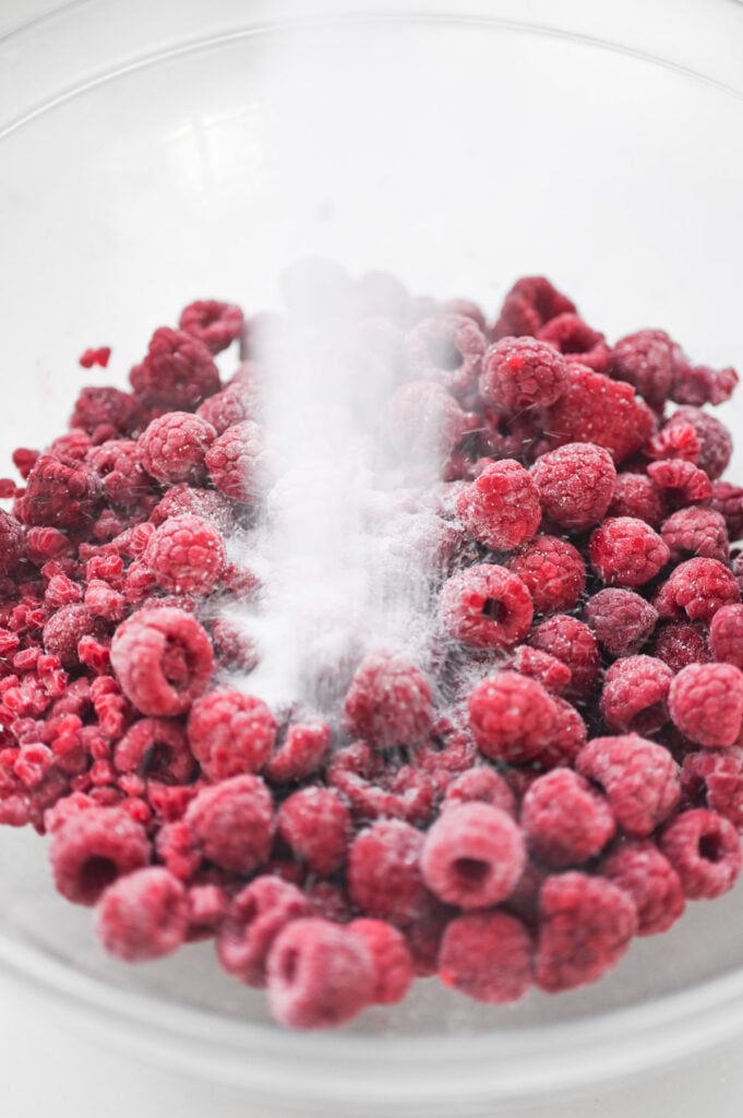 Frozen raspberries in a bowl with sugar being poured on top.
