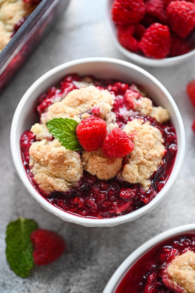 A scoop of raspberry cobbler in a white bowl with two fresh raspberries on top and a sprig of mint.