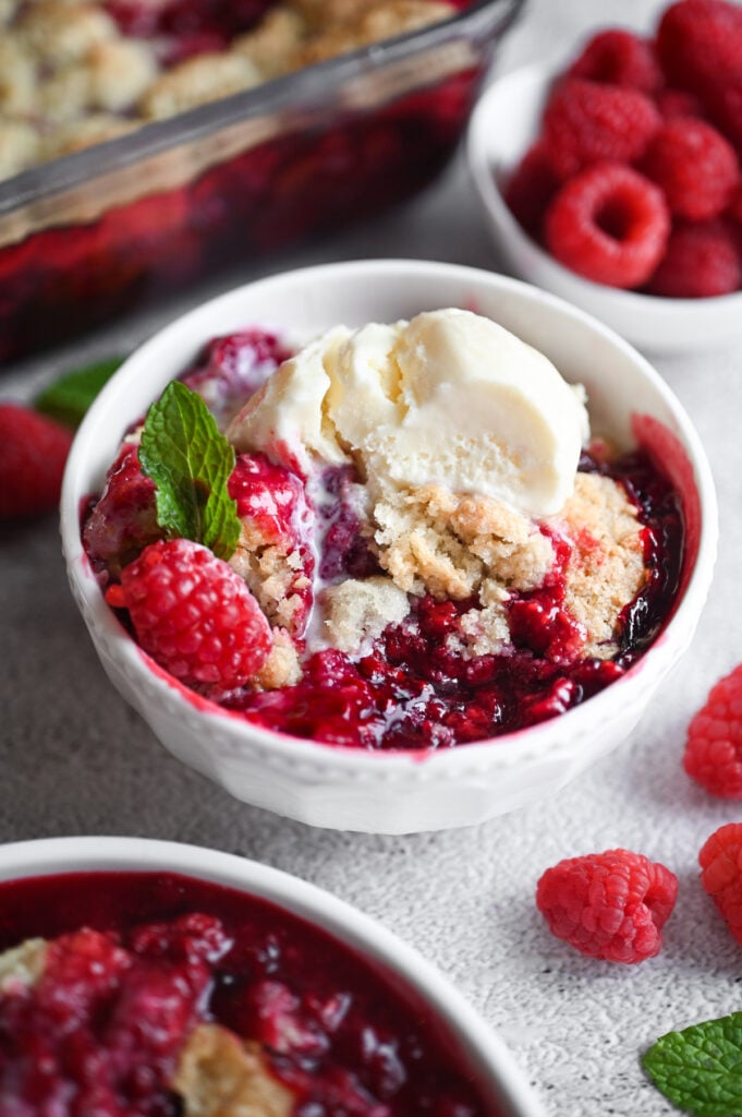 A white bowl with raspberry cobbler in it; topped with ice cream, a fresh raspberry and sprig of mint.