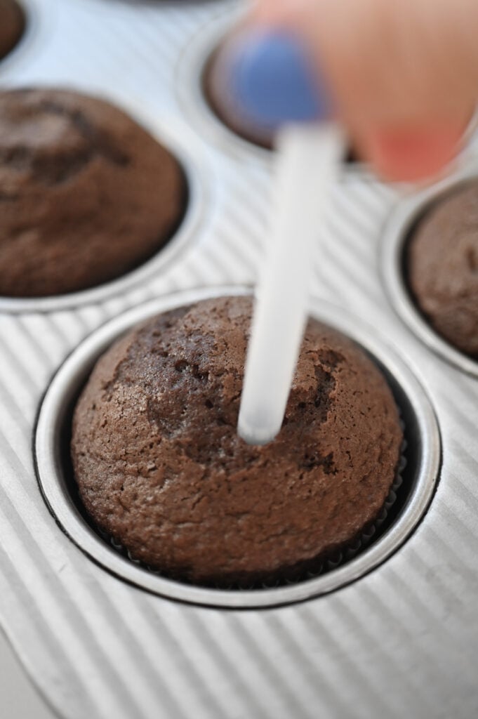 A hand holding a straw poking holes into a chocolate cupcake.