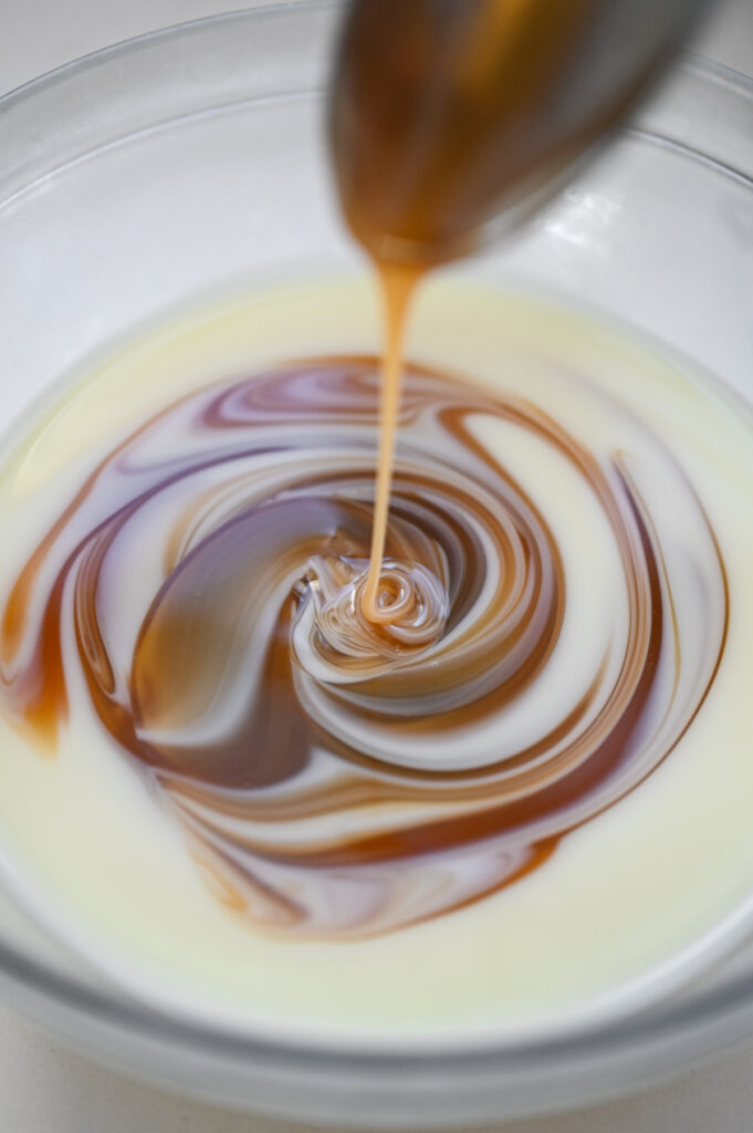 A spoon stirring caramel and sweetened condensed milk in a clear bowl.