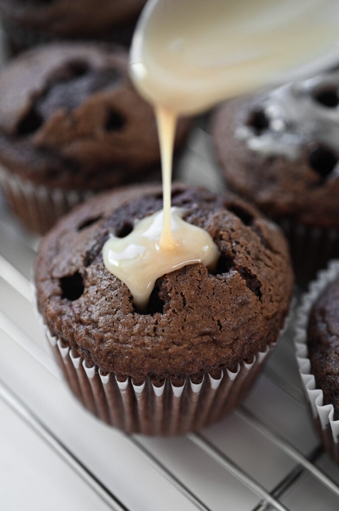 A spoon drizzling some filling into the poked holes of a chocolate cupcake.