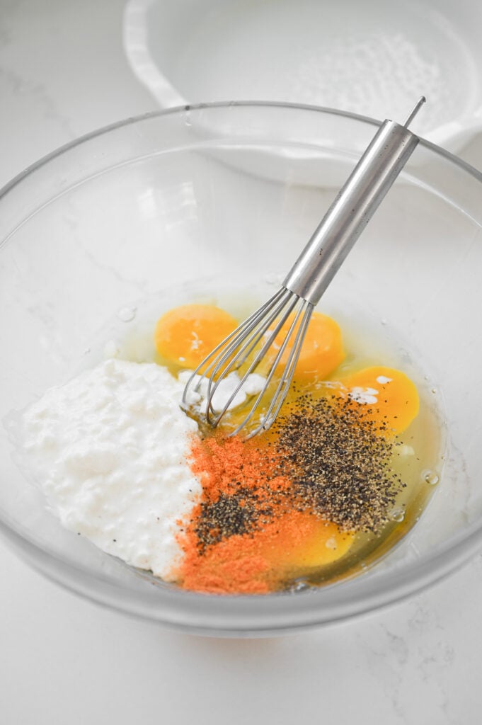 A clear glass bowl with eggs, cottage cheese, seasonings and a whisk.