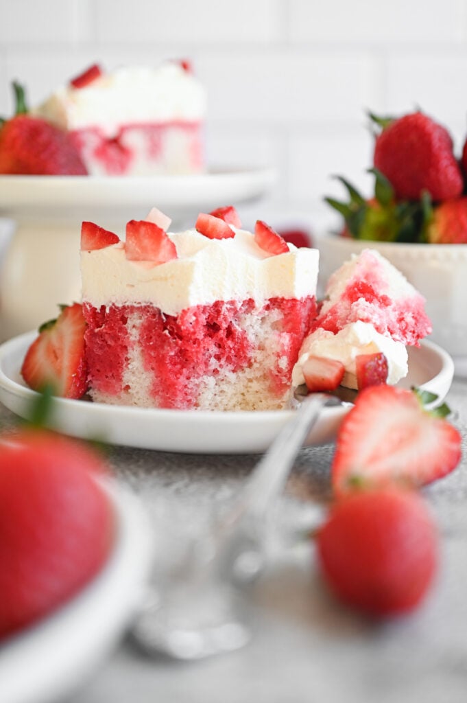 A slice of strawberry poke cake on a white plate with a couple strawberries next to it.