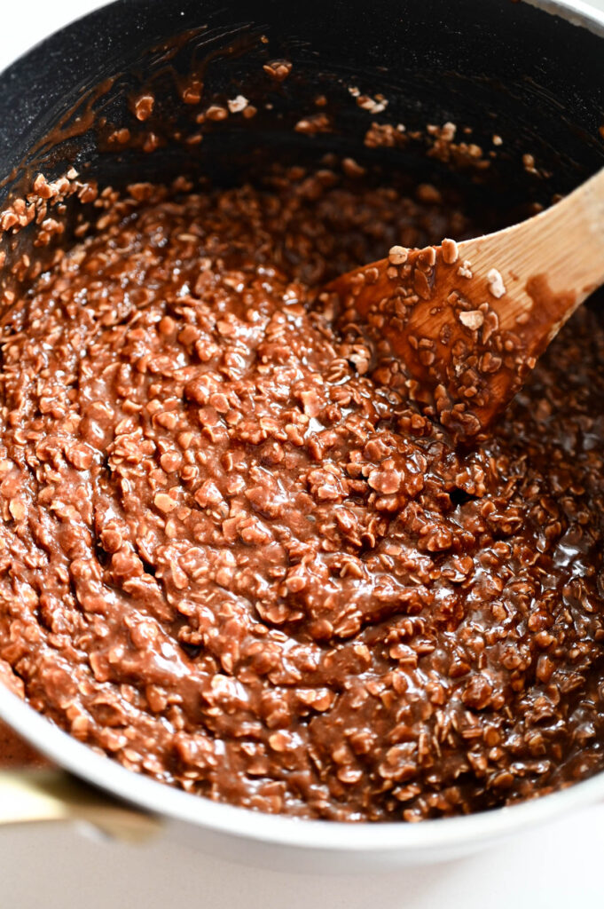A wooden spoon stirring no bake cookies in a saucepan before they're scooped.