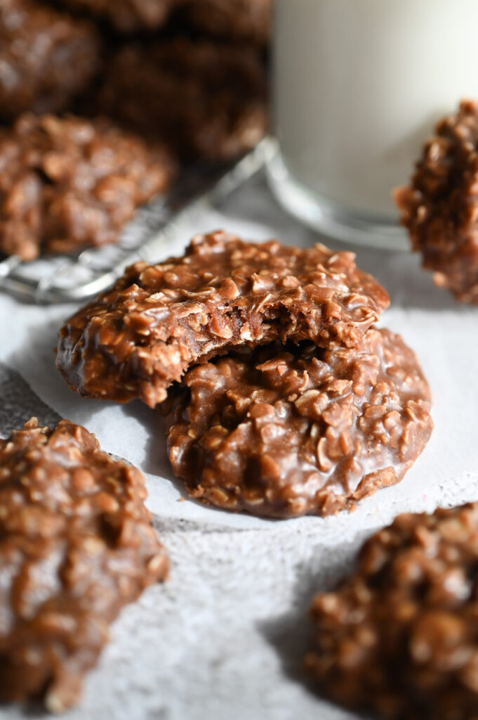 Two no bake cookies stacked next to a glass of milk; the top one has a bite out of it to show the fudgy interior.