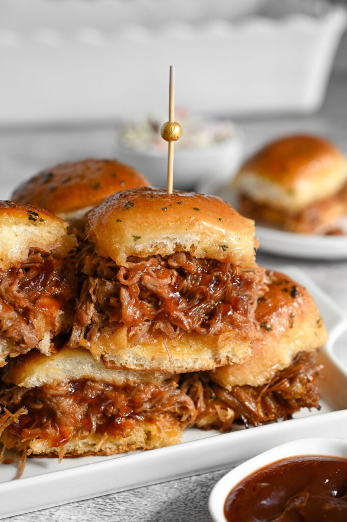 Several pulled pork sliders on a white serving tray with some bbq sauce next to it.