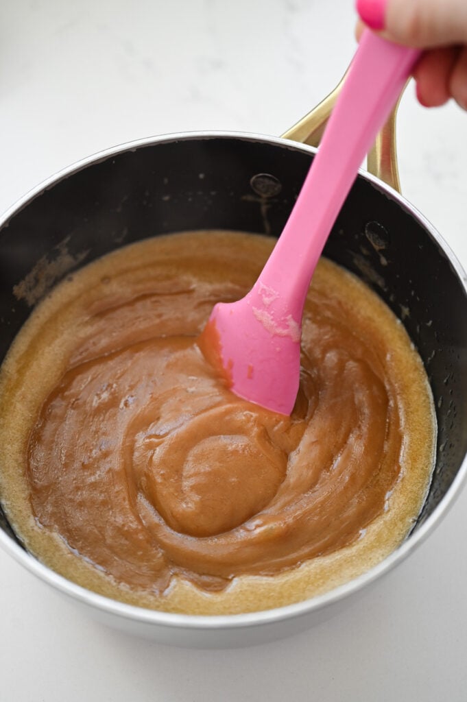 A pink rubber spatula stirring a glossy mixture with cookie butter in it.