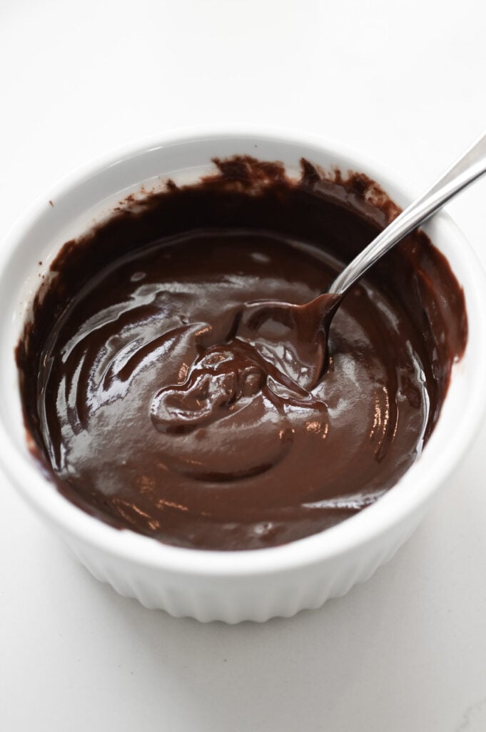 A glossy chocolate frosting being stirred in a white ceramic bowl.