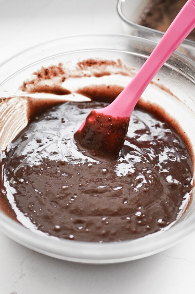 A pink rubber spatula stirring a brownie batter in a clear glass mixing bowl.