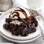 A white plate with a scoop of brownie cobbler and a scoop of vanilla ice cream on top with a chocolate drizzle.