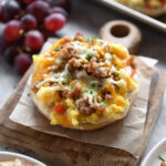 A breakfast bagel with gravy, eggs, sausage and cheese on a wooden board; grapes and a pan of more bagels in the background.