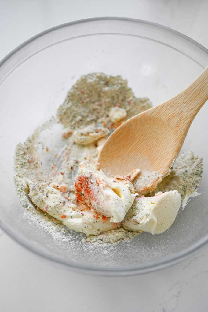 A wooden spoon stirring together cream cheese, ranch mix and other seasonings in a clear bowl.