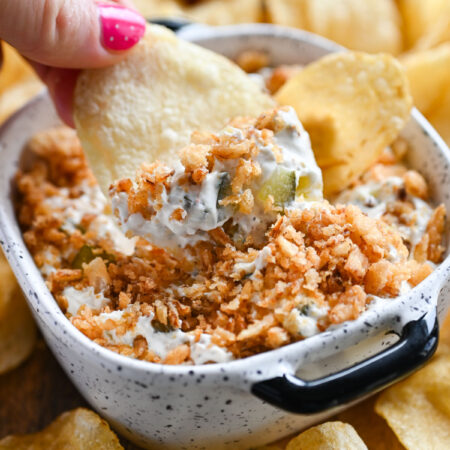A hand holding a potato chip and scooping up some fried pickle dip.
