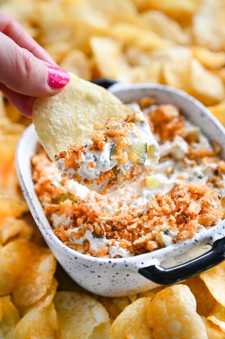 A hand holding a potato chip dipping into a bowl of fried pickle dip.