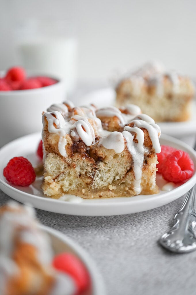 A piece of cinnamon roll casserole with frosting dripping down the side and fresh raspberries on the side, too.