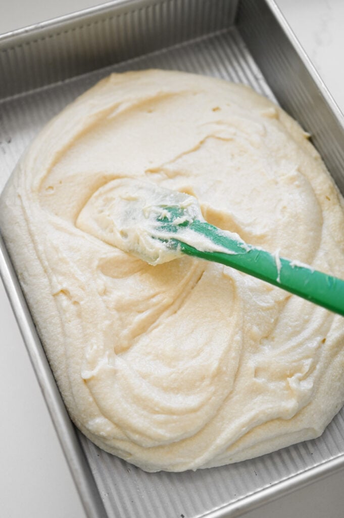 A green rubber spatula spreading cake batter out in a 9x13 inch pan.
