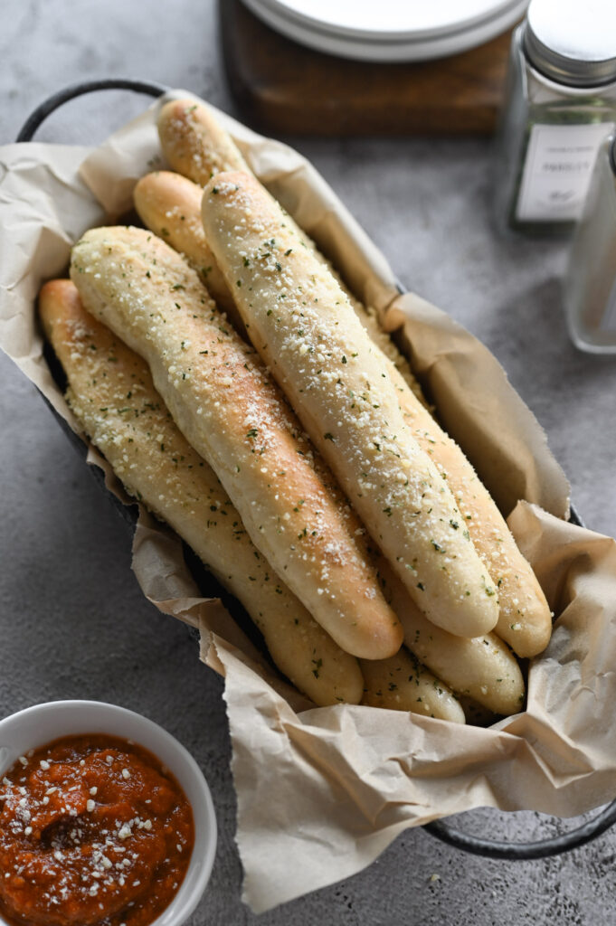 Several soft breadsticks in a basket with seasonings and a dipping sauce on the side.