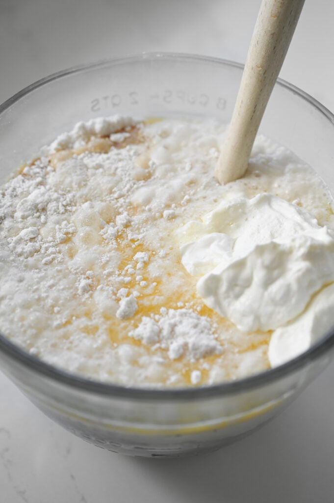 A rubber spatula in a large clear mixing bowl with many ingredients not mixed in yet.