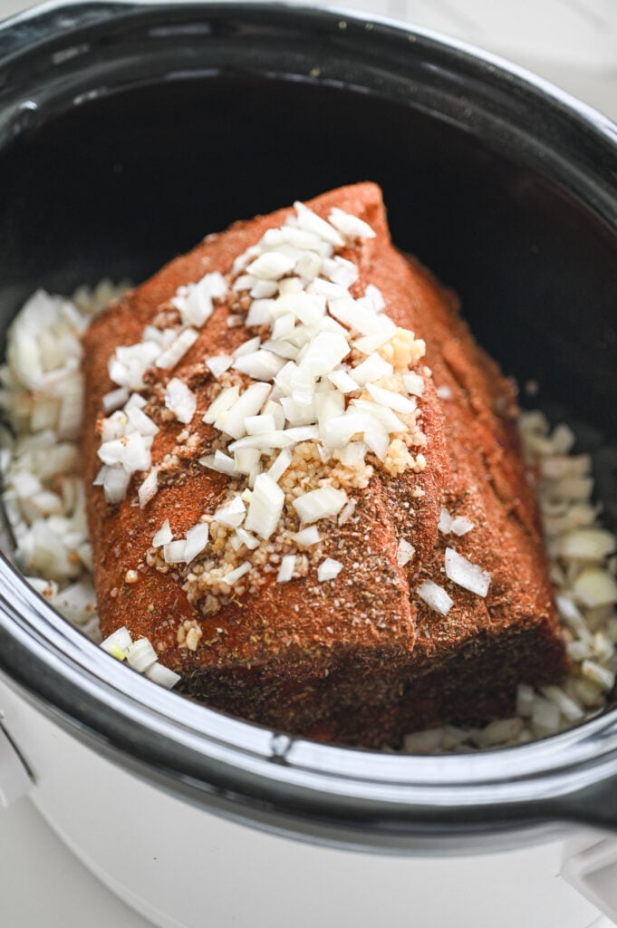 A pork roast in a slow cooker with garlic and onions on top.
