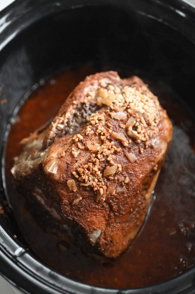 A cooked pork roast with seasonings and onions in a slow cooker.
