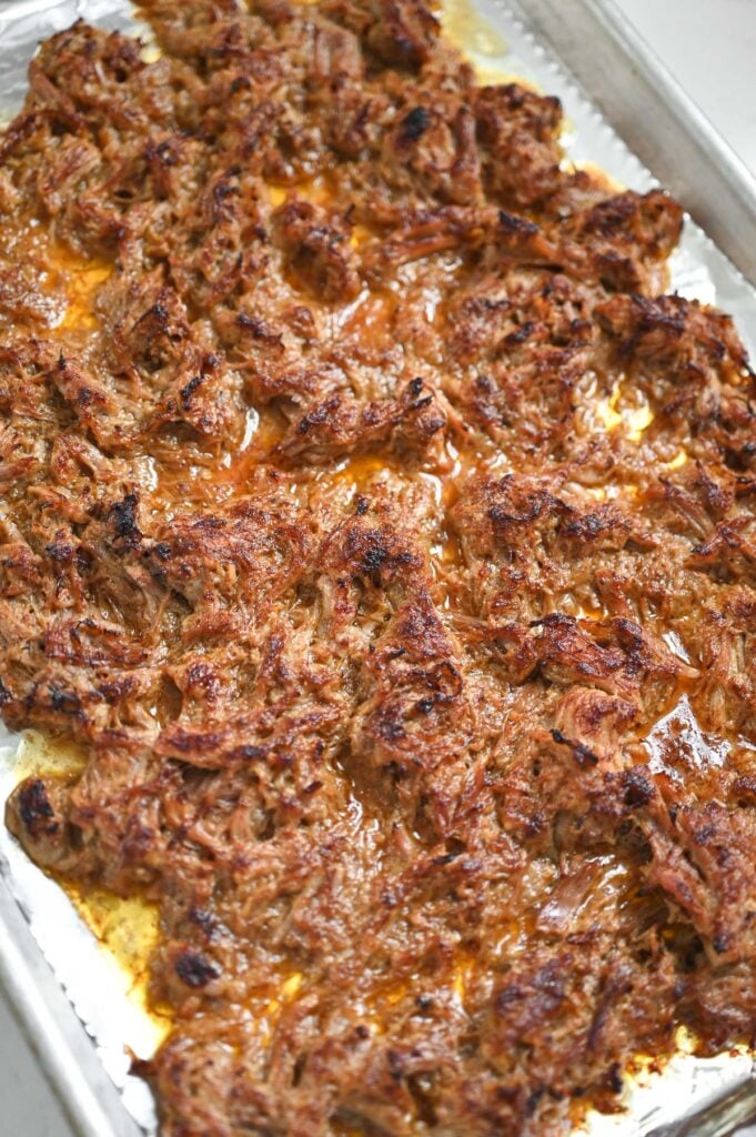 Shredded carnitas meat on a sheet pan; the tips of the meat are caramelized after being broiled.