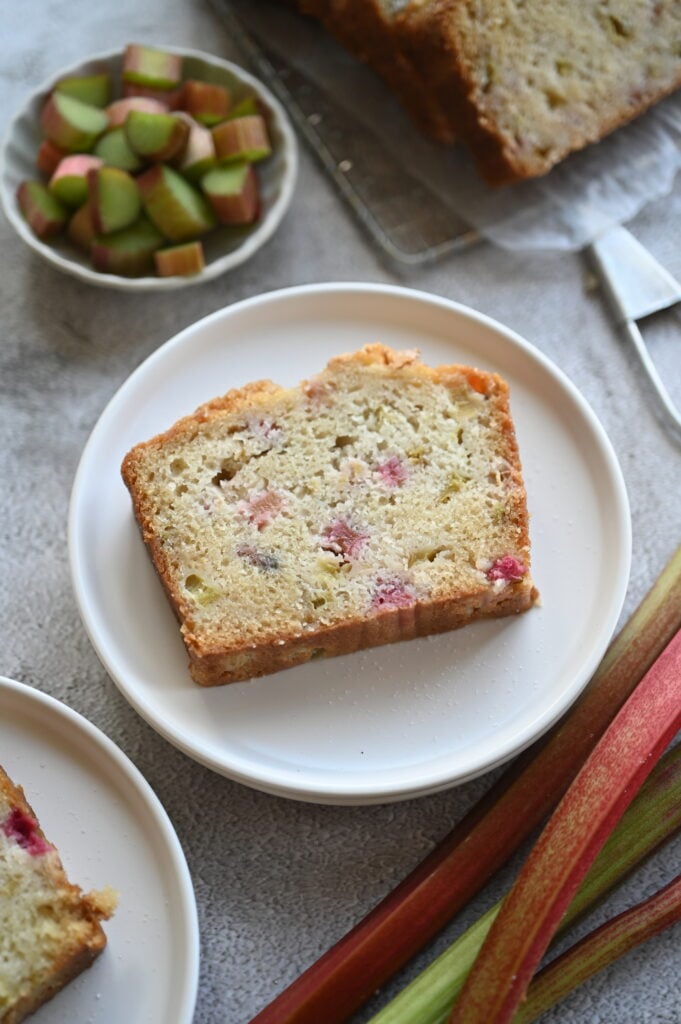 A slice of rhubarb bread on a white plate; rhubarb stalks and chopped rhubarb around it.