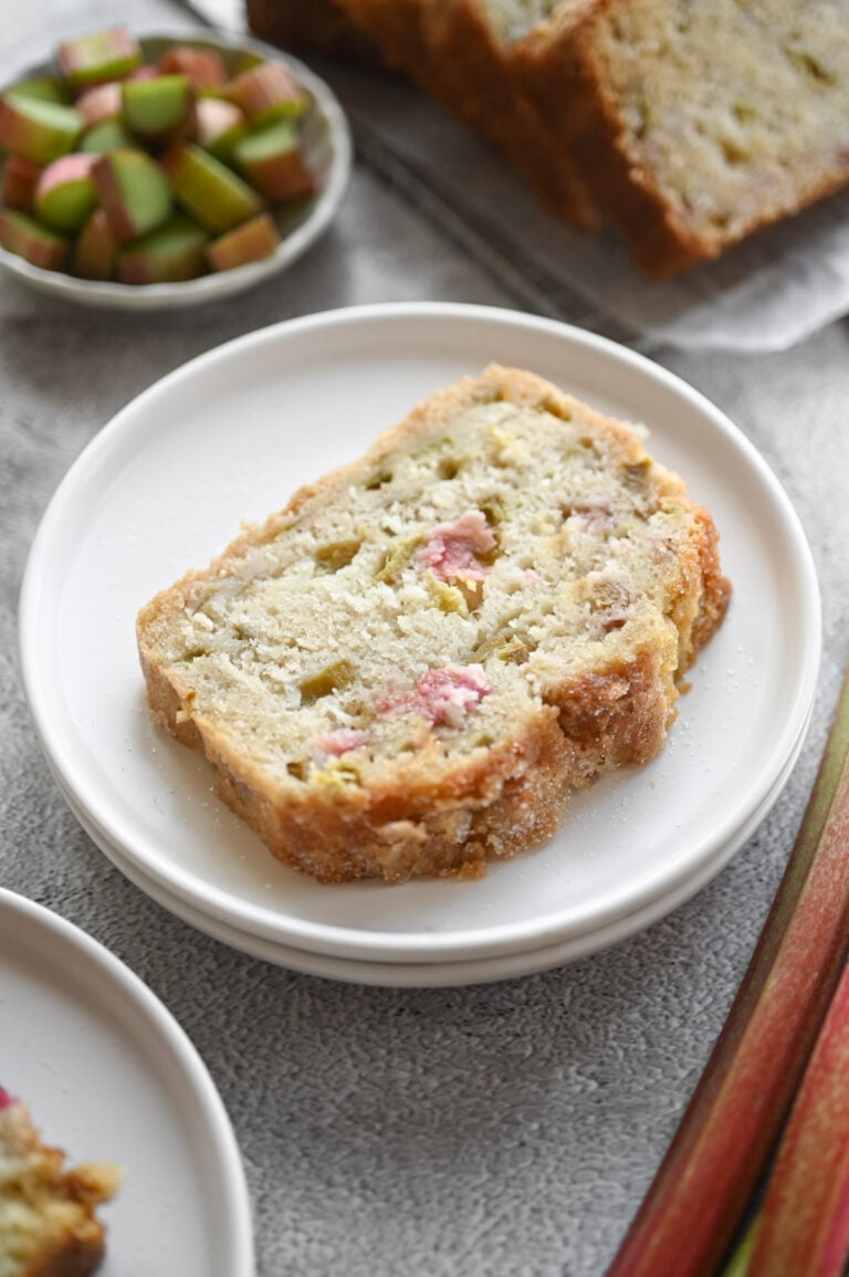 A slice of rhubarb bread on a white plate with stalks of rhubarb on the side.