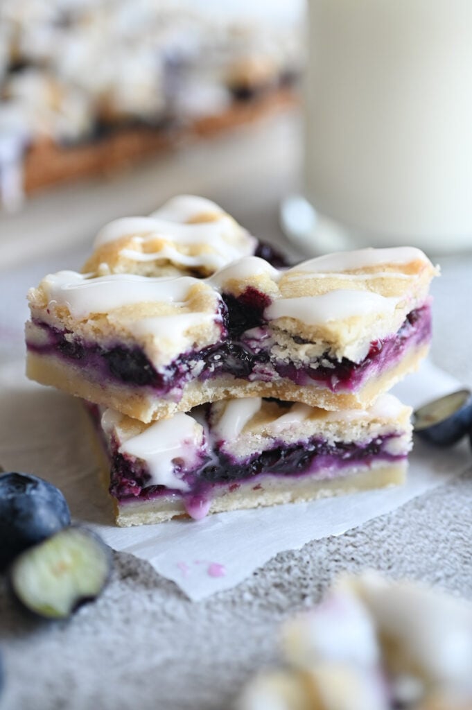 Two Blueberry Crumb Bars stacked up on a piece of parchment paper with fresh blueberries around it.