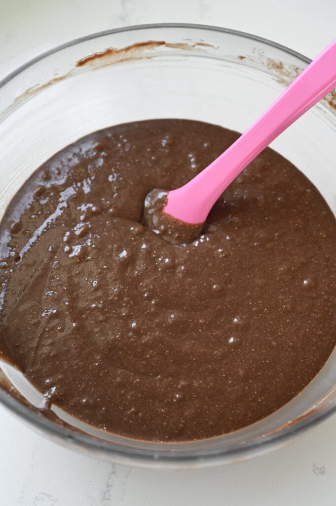 A cake batter being stirred by a pink rubber spatula in a clear glass bowl.