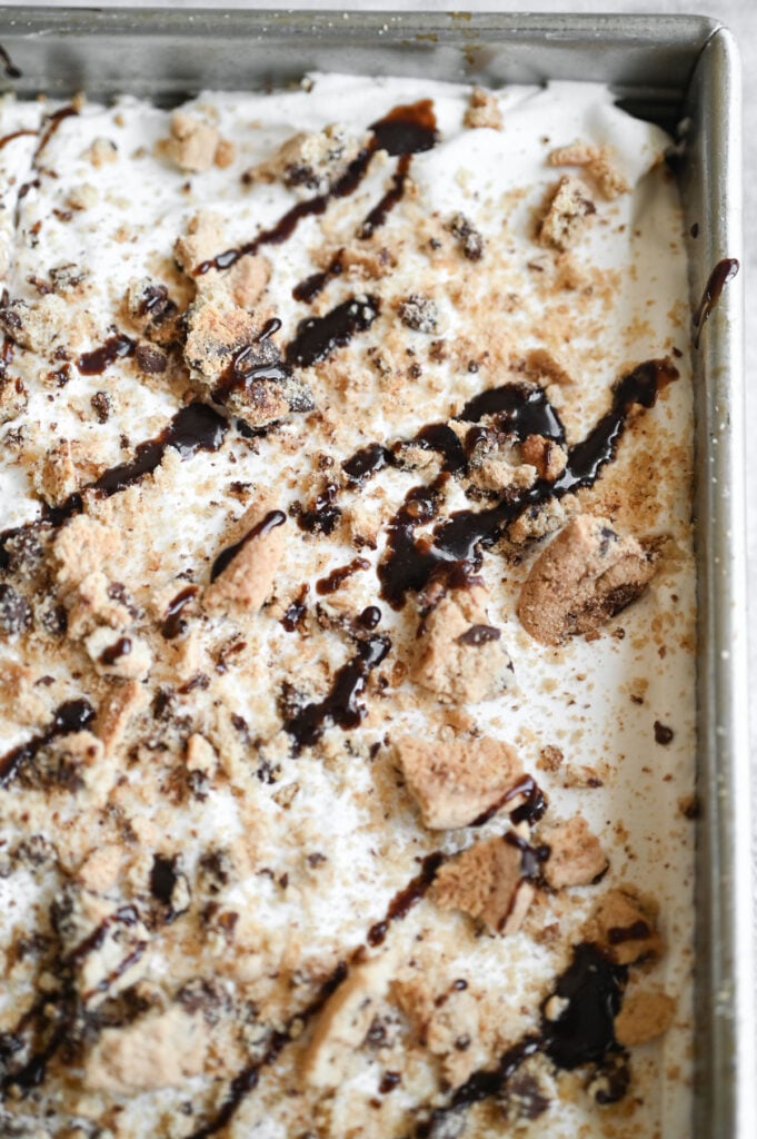 Close-up of a chocolate chip cookie poke cake topped with whipped cream, fudge sauce, and chocolate chip cookies.