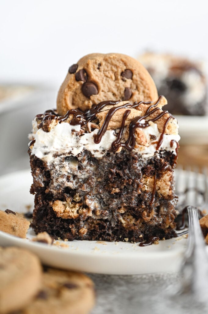 A piece of chocolate chip cookie poke cake on a white plate with a small cookie on top for garnish.