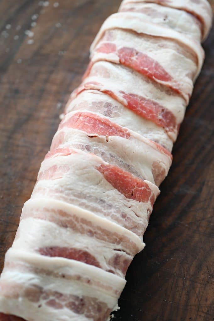 A raw bacon wrapped pork tenderloin on a wooden cutting board.