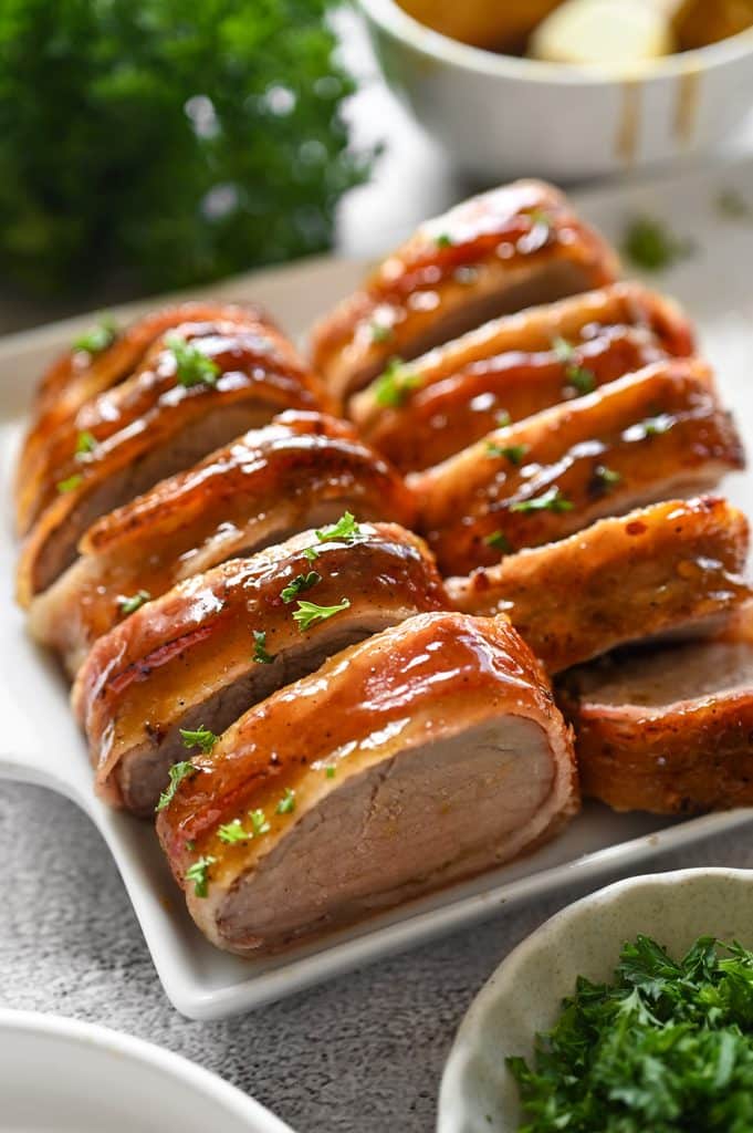 Bacon wrapped pork tenderloin cut into slices on a white platter.