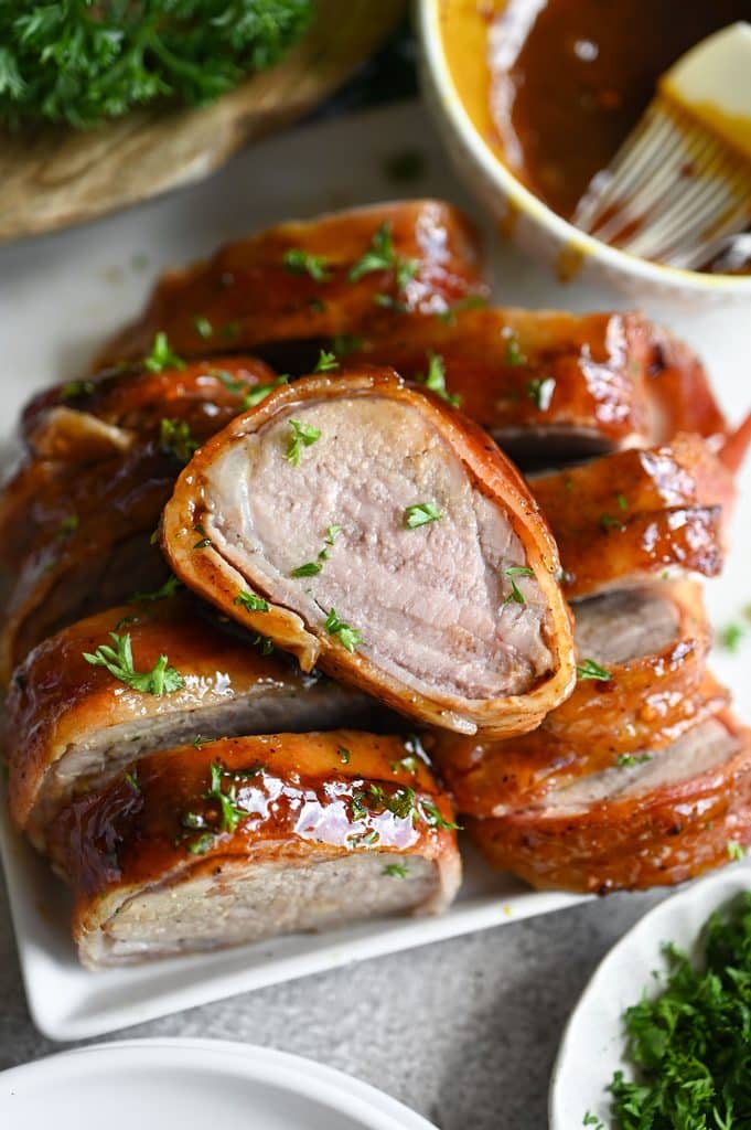 A slice of a bacon wrapped pork tenderloin on top of other slices to show the blush pink interior.
